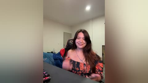 Media: Video of a young woman with long brown hair, wearing a patterned top, smiling while holding a camera, standing in a brightly lit, minimalist bedroom.