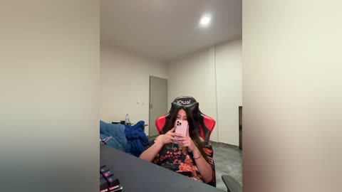 Media: Video of a young woman with long black hair in a red and black gaming chair, holding a pink smartphone, wearing a black and orange floral-patterned shirt, in a minimalist room with beige walls and a grey carpet.