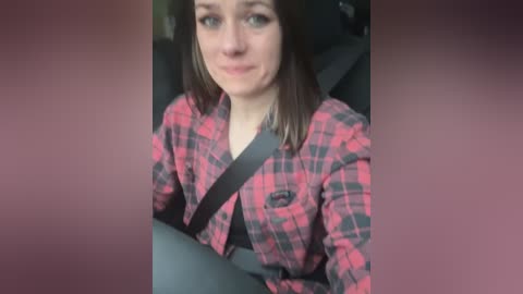 Media: A video of a woman with shoulder-length brown hair, wearing a red and black checkered flannel shirt and a seatbelt, sitting in a car seat. The background is blurred.