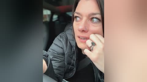 Media: Video of a woman with fair skin, brown hair, and blue eyes, wearing a black puffy jacket, looking worried, holding her face, in a car.