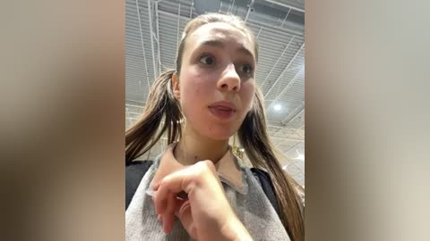 Video of a young woman with light skin, brown pigtails, and a gray shirt, taken from a low angle, showing her from the chest up. She appears to be indoors with a ceiling fan visible in the background.