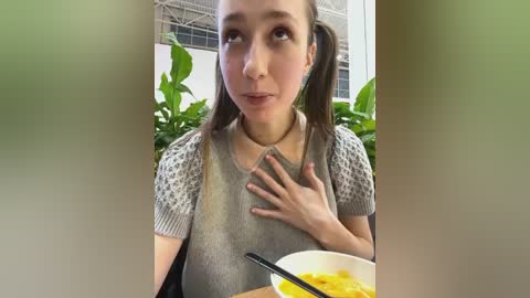 Media: A video of a young woman with light skin and pigtails, wearing a gray sweater, seated outdoors with green foliage and a white bowl of noodles in the background.