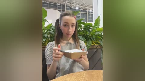 Media: Video of a young Caucasian woman with long brown hair in pigtails, wearing a light gray, short-sleeved knit top, seated indoors at a wooden table. She holds a white bowl and spoon, surrounded by lush green plants.