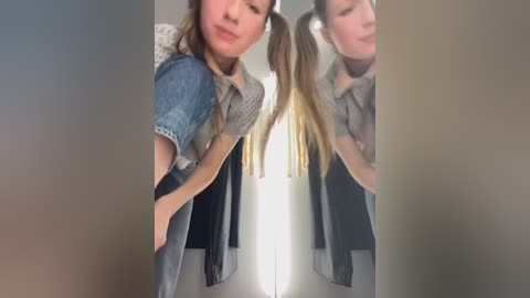 Media: Video of a young woman with long blonde pigtails, wearing a denim jacket over a grey sweater, standing in front of a mirror. She has a slight smile and is looking downward.