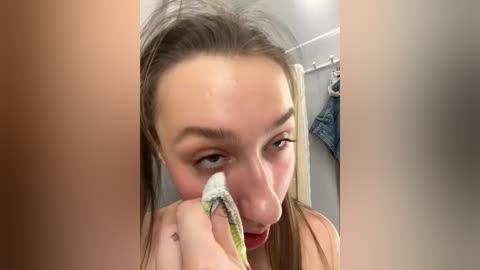 Media: A close-up video of a young woman with long brown hair, wearing a red top, wiping sweat from her face with a green and white towel in a bathroom with a hanging blue shirt.