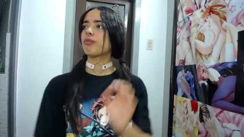 Media: Video of a young person with long, dark hair, wearing a black t-shirt with a colorful graphic, and a choker necklace, standing in a doorway with anime posters on the wall.