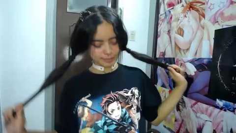 Media: Video of a young woman with long black hair, styled into twin braids, wearing a black anime t-shirt featuring \"Attack on Titan\" characters. She's in a room with anime posters on the walls.