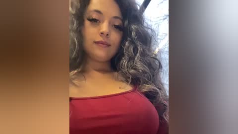 Media: A video of a young woman with long, wavy, gray-streaked hair, wearing a red off-the-shoulder top, standing indoors with a neutral background. Her skin tone is medium, and she has full lips with subtle makeup.