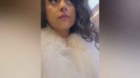 Media: Video of a woman with light brown skin, curly hair, and full lips, wearing a white fur coat, in a blurred indoor setting.