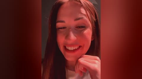 Media: Video of a young woman with fair skin, long brown hair, and light makeup, smiling with her hand near her mouth. Red lighting casts a warm glow on her face.