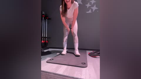 Media: Video of a fit woman with long brown hair, wearing a white tank top and leggings, performing squats on a gray mat in a gym with gray walls and a black dumbbell rack.