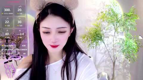 Media: A video of a young East Asian woman with long black hair, wearing a white top, and a furry headband. She has fair skin and red lipstick. Background includes a potted plant and a digital overlay of a camera screen.