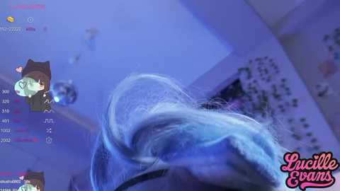 Video of a blue-haired woman in a dimly lit room, viewed from below. Text overlay reads \"Lucille Evans.\" Background includes a pink heart-shaped wall decoration.