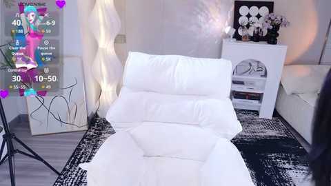 Media: Video of a modern, minimalist room with a white sofa, white pillows, and a white rug. A woman in a pink outfit is displayed on a digital overlay, with a black and white abstract wall art and a white cabinet with flowers.