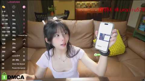 Media: Video of an Asian woman with long black hair and fair skin, sitting on a beige couch, holding a smartphone, wearing a white T-shirt and black wristband. Background shows a dimly lit living room with a kitchen visible.