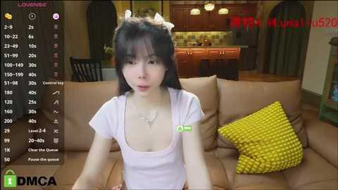 Media: A screenshot of an Asian woman with white horns, wearing a white tank top, sitting on a beige couch in a cozy living room.