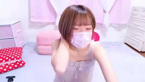 Media: Video of an Asian woman with straight brown hair and a face mask, wearing a white camisole in a pink-dressed room with white drawers.