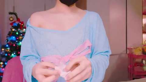 Media: Video of a light-skinned person in a blue off-shoulder sweater, holding a pink bow. Background features a Christmas tree, a pink shelf, and a black mask.