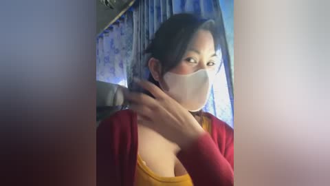 Media: Video of an Asian woman in a yellow top and red cardigan, wearing a white face mask, adjusting her hair, in a dimly lit room with blue curtains and floral wallpaper.