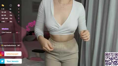 Media: A video of a woman with light skin, wearing a white cropped top and high-waisted beige pants, captured in a dimly lit bedroom.