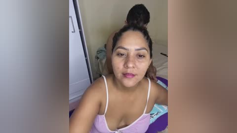 Media: A video of a Latina woman with medium skin tone and a ponytail, wearing a pink tank top, sitting on a bed with a beige wall in the background.