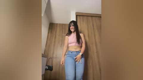 Media: Video of a smiling, light-skinned woman with long dark hair, wearing a pink crop top and high-waisted blue jeans, standing in a beige-walled room with wooden wardrobe.