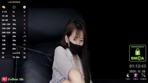 Media: A video shows an Asian woman with long black hair, wearing a black face mask, sitting on a black leather couch in a dimly lit room. The background displays a live stream with chat messages.