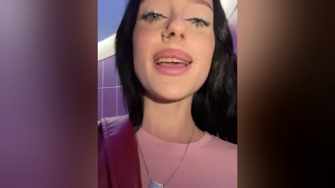 Media: Video of a young woman with fair skin, straight black hair, and a nose ring. She wears a pink top and a necklace. Background features tiled walls in purple hues.