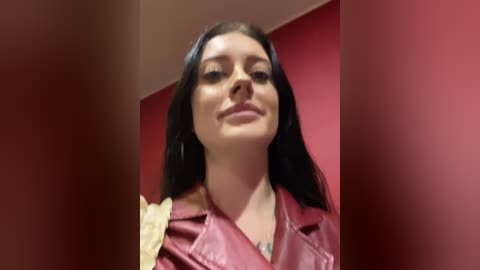 Media: Video of a young woman with long black hair, fair skin, and full lips, wearing a pink leather jacket, against a red wall.