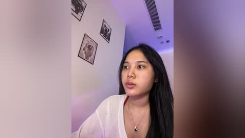 Media: Video of a young Asian woman with long black hair, wearing a white shirt, standing in a dimly lit hallway with framed black-and-white photos on the wall.