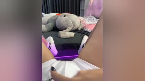 Media: Video of a person lying on a bed, wearing a black skirt and white socks, holding a purple vibrator near their crotch. The background features a gray teddy bear and a pink pillow.