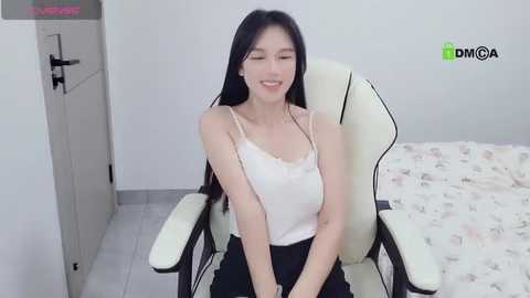 Media: Video of an East Asian woman with long black hair, wearing a white lace tank top and black pants, sitting in a white gaming chair, smiling, in a minimalist bedroom with white walls and a bed covered in floral sheets.