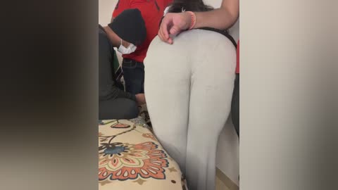 Media: A video captures a woman with a large buttocks in tight white pants, leaning against a wall, her hands gripping her waist. She wears a red top, and a man in a red shirt stands beside her.