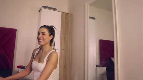 Media: Video of a smiling, fair-skinned woman with dark hair in a ponytail, wearing a white tank top, standing in a bedroom with a wooden wardrobe and red quilted bed.