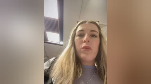 Media: Video of a young Caucasian woman with long blonde hair, wearing a light blue top and gray jacket, sitting in a bus with a window and vents in the background.