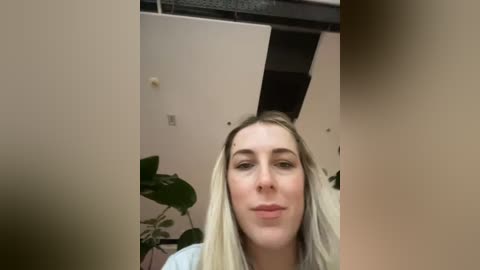 Media: Video of a fair-skinned, blonde woman with long hair, wearing a light-colored shirt, standing indoors with beige walls, a black ceiling panel, and a potted plant in the background.