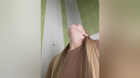 Video of a young woman with long, straight blonde hair, wearing a brown top, looking upward. She is in a green-tiled bathroom with a white wall and a brass doorknob visible.