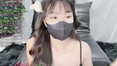 Media: Video of an Asian woman with long black hair and bangs, wearing a black face mask and a black dress, sitting on a black leather chair in a modern room with a plant in the background.