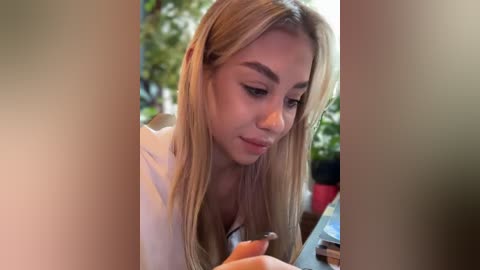 Media: Video of a young woman with long, straight blonde hair, light skin, and slender build, wearing a white top, focused on a smartphone, surrounded by blurred greenery and sunlight.