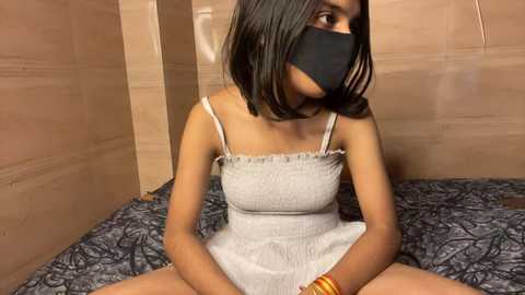 Media: Video of a young South Asian woman with medium-dark skin, wearing a black mask, light gray dress, and sitting on a bed with a dark floral-patterned sheet, against a beige wall.