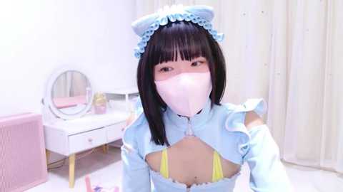 Media: Video of an Asian woman with straight black hair, wearing a light blue maid costume with a lace headband, pink face mask, and yellow lingerie. Background includes a white vanity, pink chair, and light curtains.