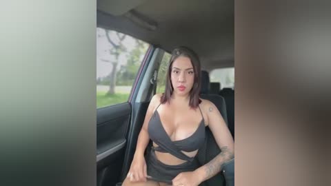 Video of a curvy, light-skinned woman with shoulder-length dark hair, wearing a black strappy top and shorts, sitting in a car, revealing a tattoo on her left arm.