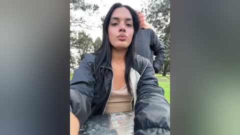 Media: Video of a young woman with long black hair, fair skin, and medium build, wearing a black and white jacket over a beige top, seated outdoors near a man in a black jacket.