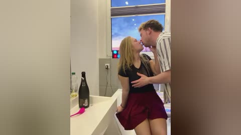 Media: Video of a blonde woman in a black top and maroon skirt, passionately kissing a man in a striped shirt at a sink in a modern bathroom.