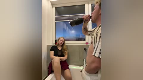 Media: Video of a blonde woman in a black top and maroon skirt sitting on a counter, watching a shirtless man drinking from a bottle in a bathroom with a window showing a cloudy sky.