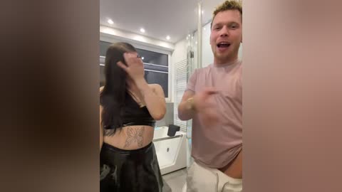 Media: Video of a woman in a black latex outfit and a man in a light pink shirt and white pants in a modern bathroom with white tiles and a bathtub.