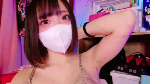 Media: Video of an East Asian woman with straight black hair and blunt bangs, wearing a white face mask, black bra, and a black wristband. She's in a cluttered room with a pink curtain, a wooden shelf, and various items.