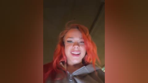 Media: Video of a smiling, fair-skinned woman with vibrant, shoulder-length red hair, wearing a gray hoodie. The background is dimly lit with a blurry, warm tone.