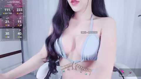 Media: Video of a light-skinned woman with long, wavy dark hair in a blue bikini, standing indoors with a calendar and digital clock in the background.