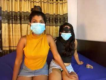 Media: Video of two South Asian women, one with a dark complexion and dark hair in a bun, wearing a yellow top and denim shorts, and the other with a lighter complexion and long dark hair, wearing a black top and checkered shorts. Both wear blue masks. They sit on a blue bed with beige and black curtains in the background.